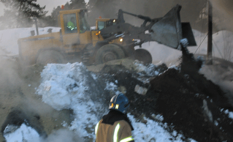 Ti ansatte ved avfallsanlegget hjelper brannmannskapene, ved å dumpe store mengder jord og sand over flammene. Foto: Marianne Aakermann.